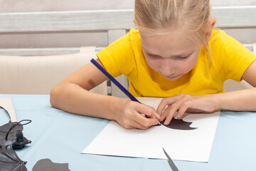Naklejka premium A girl draws and cuts a Halloween decoration at home from black paper. DIY craft decorations. Child makes black bats