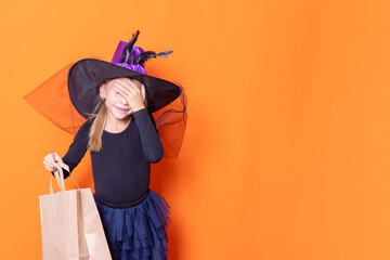 A beautiful girl in a black dress and a witch hat holds a craft paper bag, covering her face with her hand, wondering