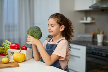 Fototapeta premium Front portrait of a little girl with curly hair, holding and looking at broccoli, seated at table in the kitchen.