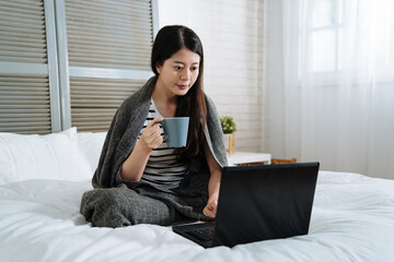 Happy casual beautiful asian korean woman working on laptop computer sitting on white bed in bright house. elegant female cover body with warm blanket and enjoy morning hot coffee in winter bedroom