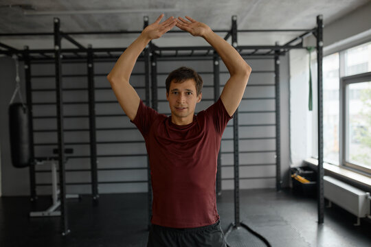 Horizontal image of good-looking middle-aged male in red fitness shirt warming up putting hands up together looking straight at camera isolated on background of modern gym, relieving stress