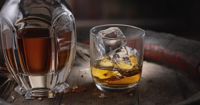 An ice cube slowly falls into a glass of whiskey on top of a vintage whiskey barrel. A decanter with a drink stands nearby, a dark brown background. 