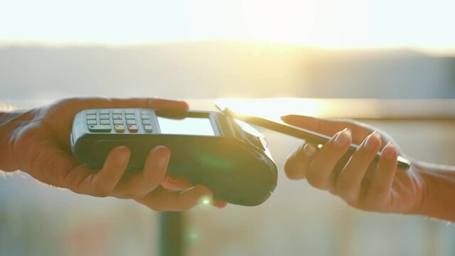 Close-up, woman using smartphone cashless wallet NFC technology to pay order on bank terminal. Card machine in male hand on sunlight background