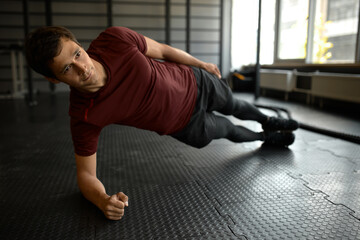 Horizontal side view of cheerful man doing side plank exercise on elbow with ease, looking calm and confident, wearing stylish sportswear and sneakers, training at gym. Abs workout