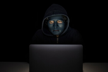 Hacker with face mask working on laptop computer in dark room