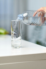 Female hand holding drinking water bottle and pouring water into glass on table.
