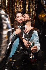 Fototapeta na wymiar joyful and stylish couple clinking champagne glasses near sparkling christmas decoration on black