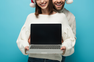 partial view of smiling couple in santa hats holding laptop with blank screen isolated on blue