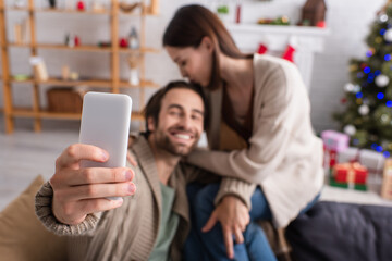 selective focus of smartphone in hand of happy man taking selfie with wife in decorated living room