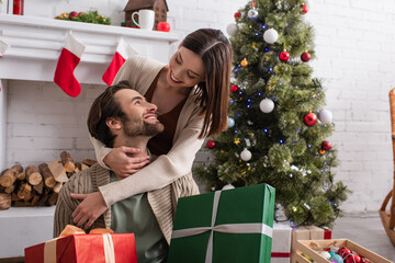 pleased woman embracing husband holding gift boxes near fireplace and christmas tree