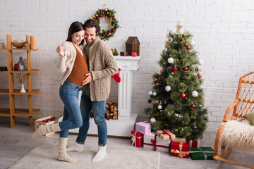 full length view of pregnant woman with happy husband near fireplace and christmas tree in living room