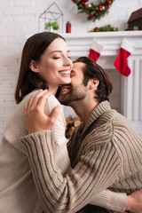 pleased man hugging wife near blurred fireplace with christmas decoration