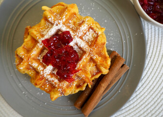 on a gray plate are Belgian gluten-free pumpkin waffles, sprinkled with powdered sugar and red currant jam, with cinnamon sticks. top view