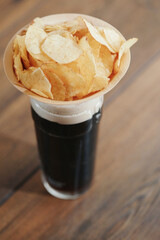 Dark ale in a glass with foam and a wooden small cup with potato chips on a wooden table. Selective focus. Traditional beer and snack