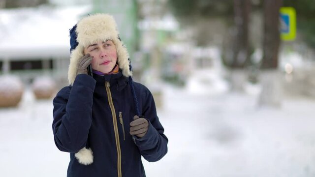 Portrait of a young woman in a hat with earflaps and a jacket, who calls and talks on the phone. Snow-covered street in the background.