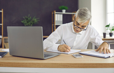 Senior businessman doing paperwork and taking notes in his business planner or notebook. Busy older adult man sitting at office desk with modern laptop and writing something down on sheet of paper