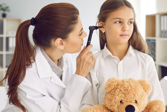Little girl with teddy bear getting ENT examination in exam room at routine checkup at doctor's office. Female otolaryngologist holding otoscope and examining child's ear before prescribing therapy