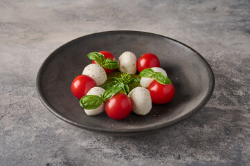 Delicious italian caprese salad with ripe tomatoes, fresh basil and mozzarella cheese in black plate on the background as concrete. Close up 