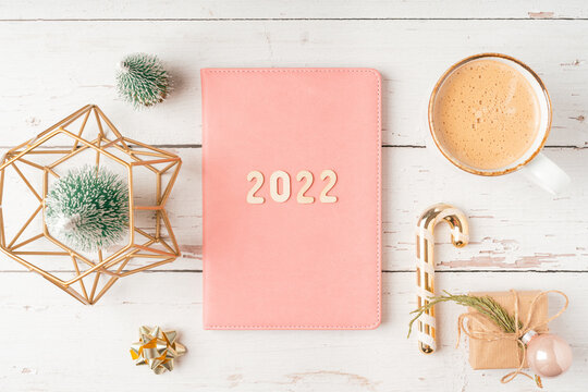 Top view Desktop Christmas pink notepad with 2022 letters text. Flat lay on white wooden table background with planner, cup of coffee, candle, toys, Christmas decoration, notebook and stationery.