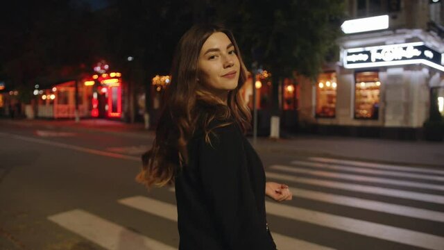 Trendy young girl goes along a pedestrian crossing in the evening and heads to a summer cafe illuminated with neon lights and colorful garlands