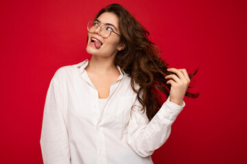 Beautiful young happy joyful funny curly brunette woman wearing white shirt and optical glasses isolated on red background with copy space
