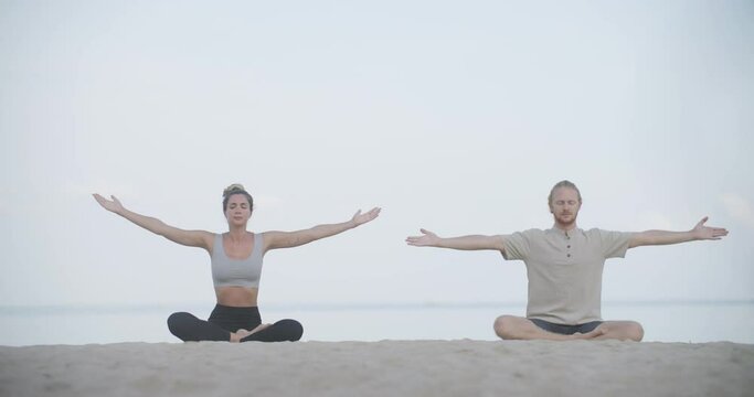 Young and healthy caucasian man and woman sitting on the beach and practicing amazing yoga lotus pose together for relaxing in the morning. Ocean in background. Health, Wellness and Meditation Concept