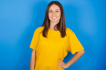 Studio shot of cheerful Young european brunette woman wearing yellow T-shirt on blue background...