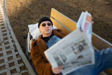 A lying man on a bench is reading a newspaper outdoors.