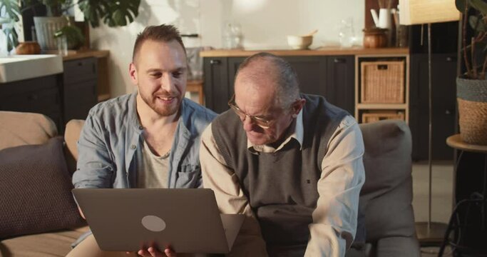 Helping elderly people to use modern technology. Happy young adult man teaching 70s senior dad to use laptop at home.