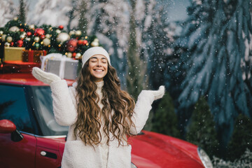 Happy beautiful woman wearing knitted sweater and woolen hat standing near red car with Christmas...