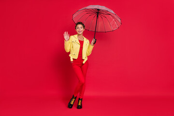 Photo of excited friendly young woman dressed leather jacket holding open parasol walking waving arm smiling isolated red color background