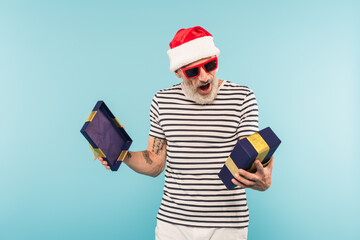 amazed middle aged man in santa hat and sunglasses opening gift box isolated on blue