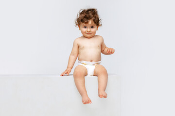 Portrait of little cute toddler boy, baby in diaper joyfully sitting isolated over white studio...