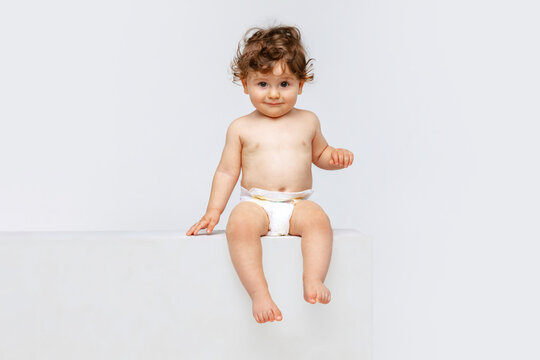 Portrait of little cute toddler boy, baby in diaper joyfully sitting isolated over white studio background. Happy childhood