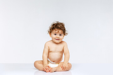 Portrait of little cute toddler boy, baby in diaper joyfully sitting isolated over white studio background. Happy childhood - obrazy, fototapety, plakaty
