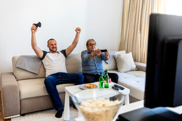 Smiling senior dad and his excited son are sitting on the sofa in the living room and enjoying time together while playing console games with game controllers. Family having fun at home.