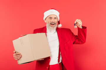 Fototapeta na wymiar excited middle aged man in suit and santa hat holding carton box and key isolated on red