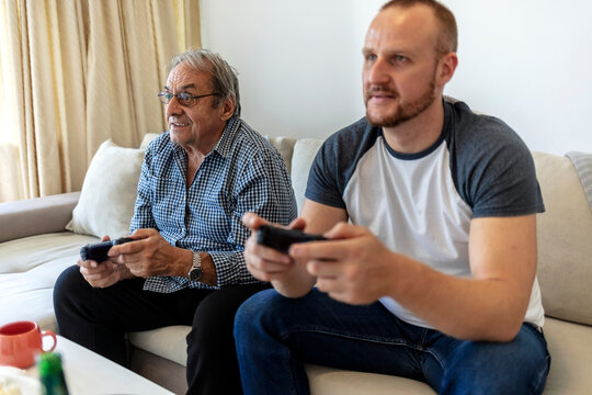 Father and his son spending their free time playing video games and enjoying each others company. Overjoyed senior father sit relax on couch with son playing computer game with joysticks.