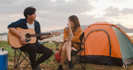 Young asia campers couple playing guitar serenading each other near beach. Male and female travel happy romantic moment when sunset in evening. Outdoor activity, adventure travel, or holiday vacation.