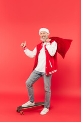full length of cheerful middle aged man in hat holding santa sack while riding skateboard and...