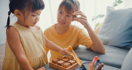 Happy cheerful Asia family mom teach girl play board game hobby with wooden box having fun relax on couch in living room at house. Spending time together, Social distance, Quarantine for coronavirus.