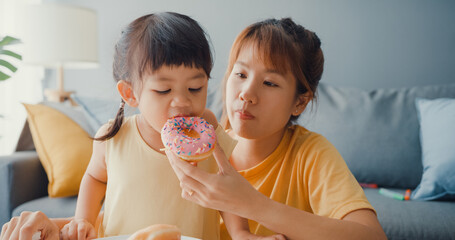 Happy cheerful Asia family mom and toddler girl eating donuts and having fun relax enjoy on couch...