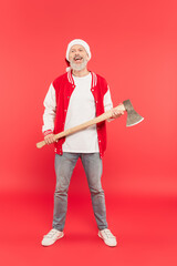 full length of smiling middle aged man in santa hat holding axe on red