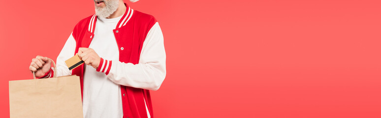 cropped view of middle aged man putting credit card in shopping bag isolated on red, banner