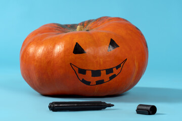 Halloween pumpkin with drawing Jack-o-Lantern face prepared for cutting. Blue background, black marker lying down near, holiday concept. Copy space.