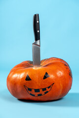 Halloween pumpkin with drawing Jack-o-lantern face and kitchen knife sticking out from above, on blue background. Copy space, holiday preparation concept.