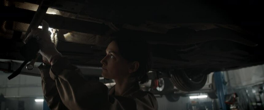 Caucasian female mechanic repairing a car in a workshop, working under car bottom. Shot with 2x anamorphic lens