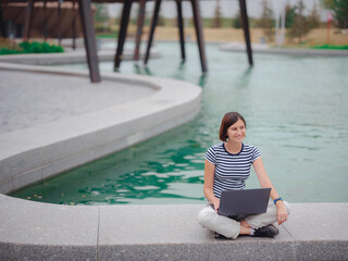 happy asian female student working with laptop in modern university campus park. woman in striped T-shirt and light trousers typing at gray laptop against the backdrop of a pond.