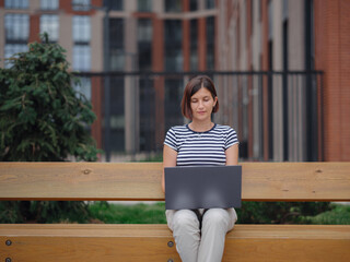 Young asian female student working at laptop sit down on bench outside on urban city street. Happy lady distance learning, education and online shopping.
