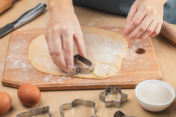 The hand holds a metal mold and cuts out heart-shaped cookies from the dough. Top view. Wooden cutting board, flour,eggs, knife, sugar. eggs.